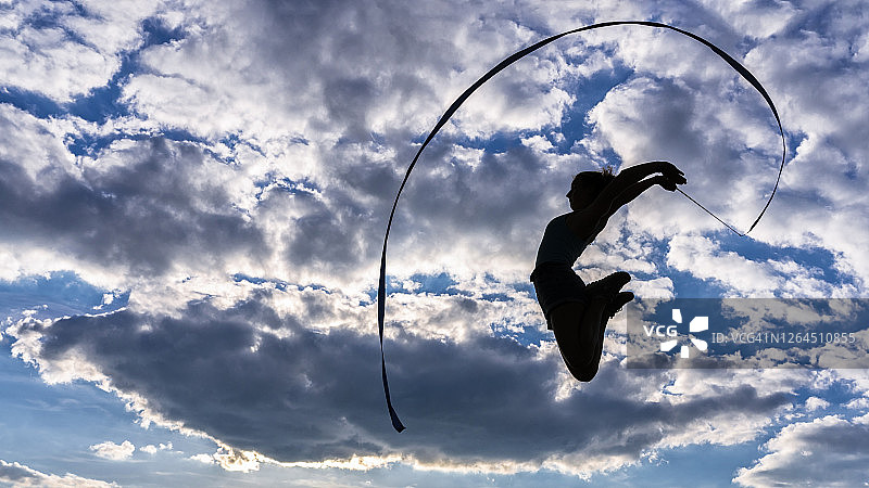 年轻女子拿着彩带跳跃，在多云的天空下做健美操图片素材
