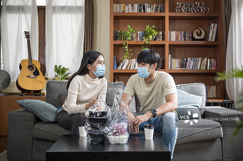 年轻夫妇戴着口罩坐在沙发上，在客厅里一起用餐图片素材