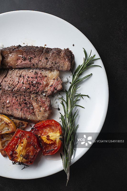 白盘盛放的烤和牛牛排切片配烤柠檬、番茄、大蒜和迷迭香图片素材