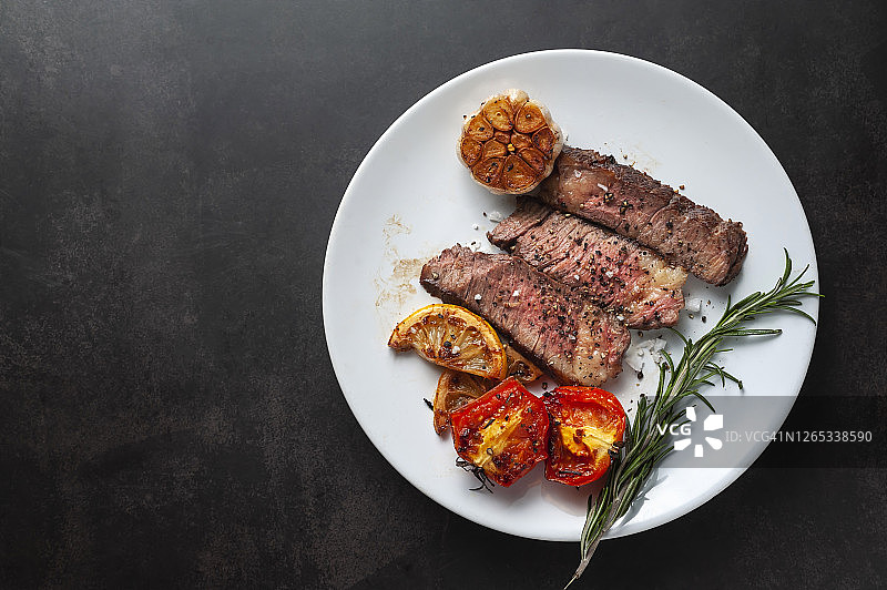 白盘盛放的烤和牛牛排切片配烤柠檬、番茄、大蒜和迷迭香图片素材