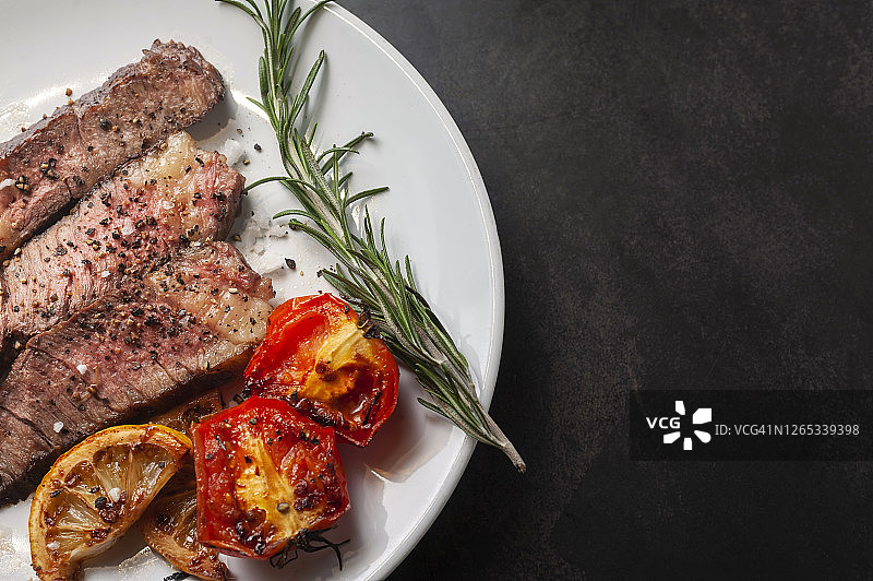白盘盛放的烤和牛牛排切片配烤柠檬、番茄、大蒜和迷迭香图片素材