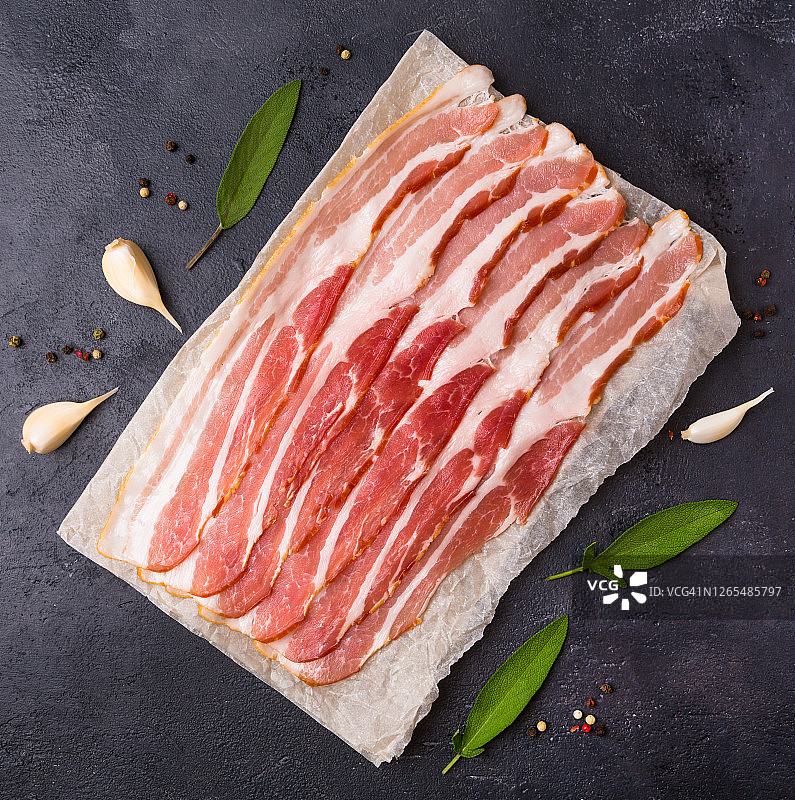 黑色背景上待烹饪的生培根片，配有调味料和香草，以及可直接食用的意式火腿片。图片素材
