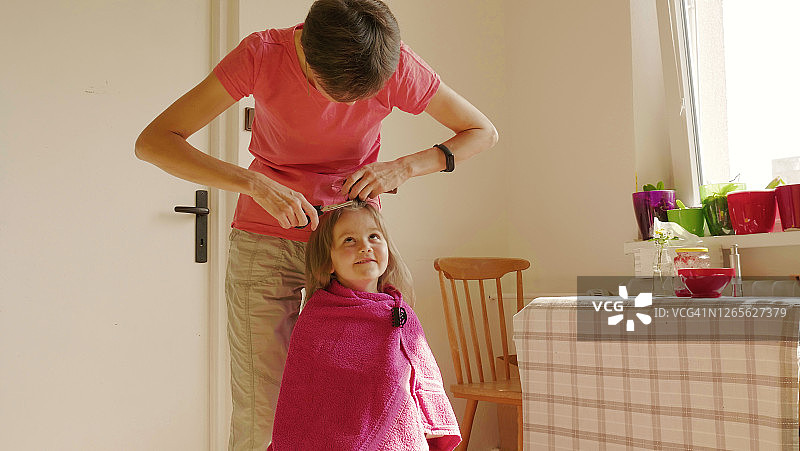 疫情隔离期间，在家中厨房理发的母亲看着微笑的金发小女儿图片素材