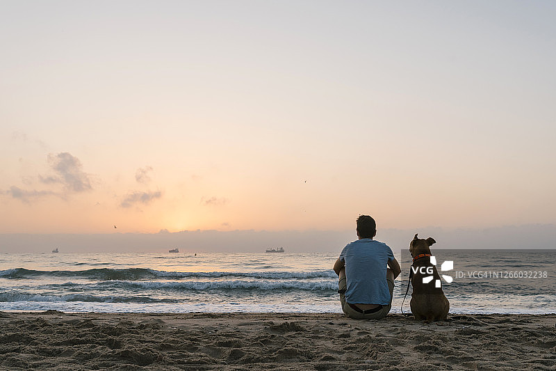 黎明时分，男子和他的狗坐在沙滩上图片素材