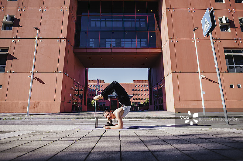 年轻女子在城市街道的建筑物旁，通过笔记本电脑练习前臂倒立图片素材