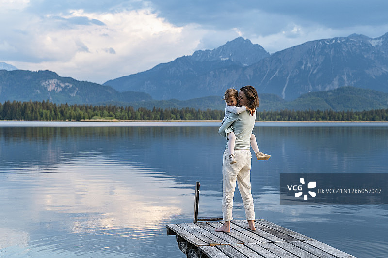 快乐的母亲扛着女儿站在湖边的码头上，背景是山图片素材