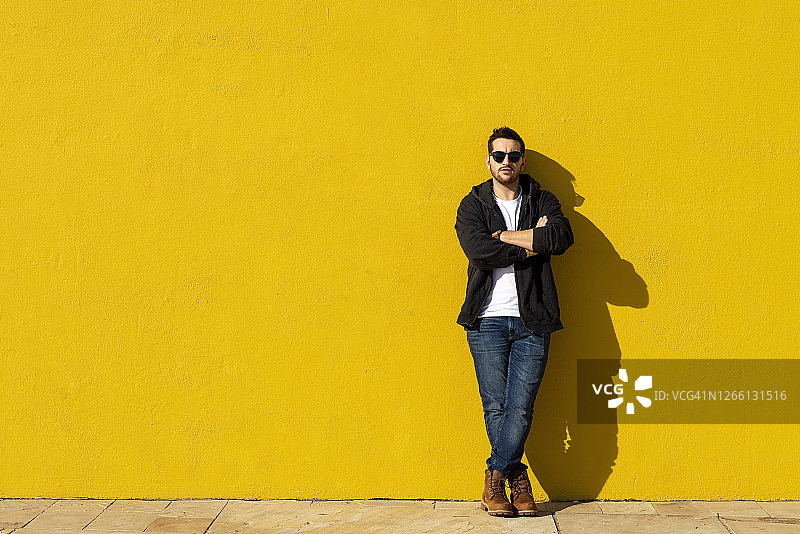 双手交叉靠在黄色墙壁上的年轻络腮胡男子图片素材