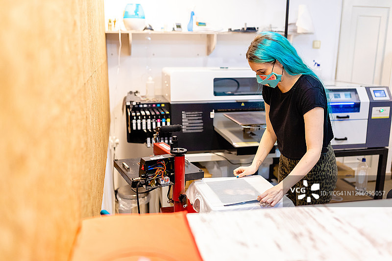 在数码印刷车间，女工在新冠疫情期间戴着防护口罩准备印刷T恤图片素材