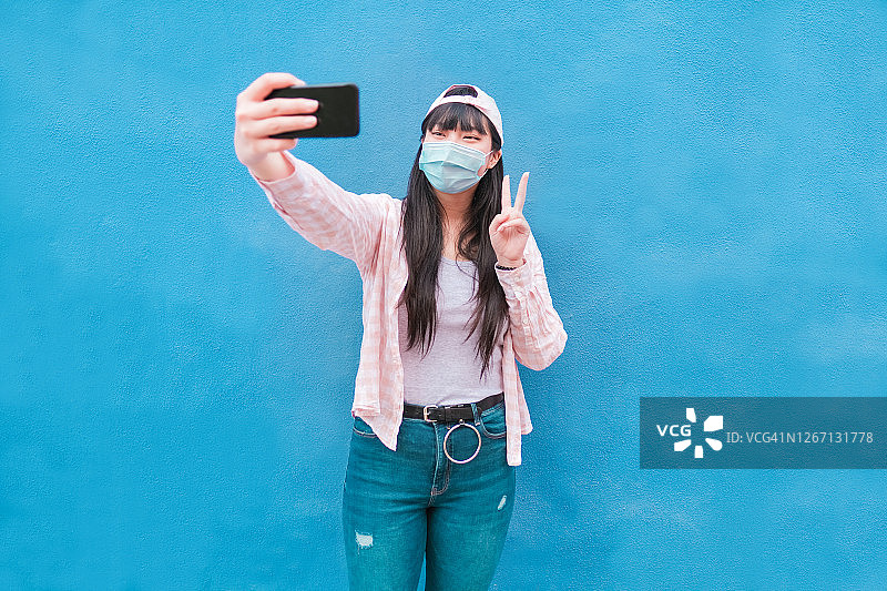 快乐的亚洲女人戴着防护口罩，用智能手机自拍，同时用手做出和平标志 - 专注于面部 - 冠状病毒生活方式图片素材