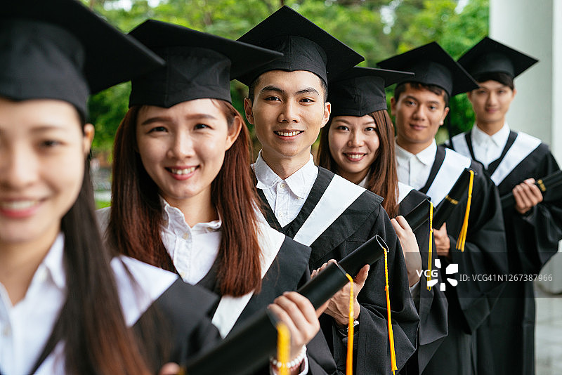 穿着毕业礼服，拿着文凭，站着表达快乐的亚洲学生图片素材