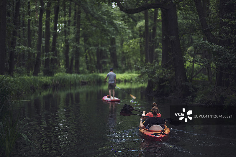 在德国勃兰登堡州施普雷瓦尔德吕贝瑙，两名女子乘坐皮划艇，一名男子站在桨板上漂浮在森林中图片素材