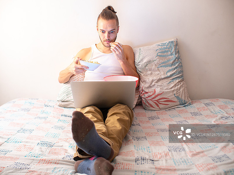 白人男子在床上用笔记本电脑看电影吃爆米花图片素材