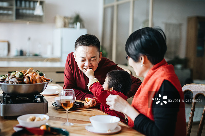 快乐的亚洲多代同堂家庭一起庆祝中国新年，可爱的小孙子在团圆晚宴上与祖父分享美味的传统中国盆菜图片素材