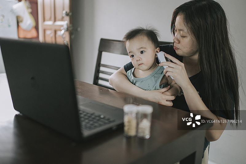 远程医疗：中国台湾母亲用体温计为宝宝测量体温，同时使用笔记本电脑在线咨询儿科医生图片素材