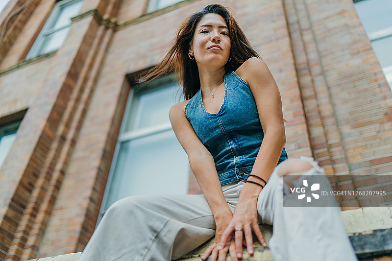 年轻女子以酷酷的姿态坐在砖墙前的仰视肖像图片素材