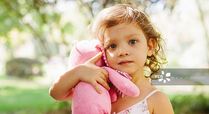 在公园里抱着洋娃娃在树旁玩耍的可爱小女孩图片素材