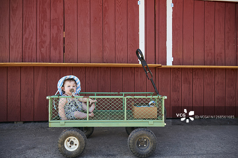 戴着遮阳帽坐在红色谷仓背景的婴儿车里的宝宝图片素材