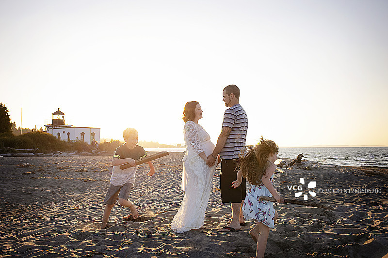 海滩上，丈夫和怀孕的妻子手牵着手，孩子们在他们周围玩耍图片素材