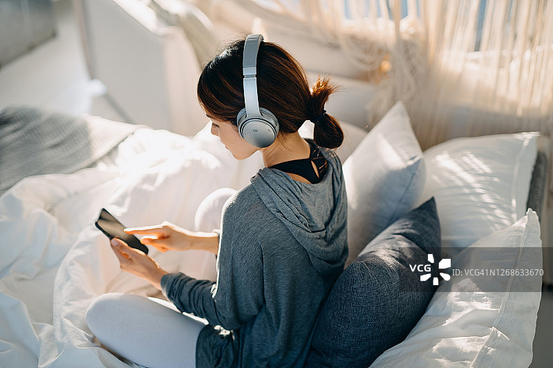 在床上戴着耳机听音乐，用智能手机上网的放松的年轻亚洲女人图片素材