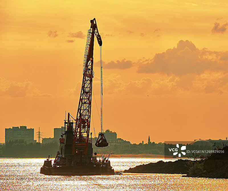 在岸边附近工作的疏浚起重机，背后的傍晚橙色阳光反射图片素材