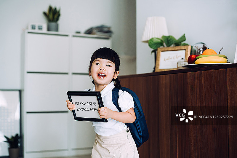 快乐的亚洲小女孩穿着校服，拿着“幼儿园第一天”的标牌图片素材