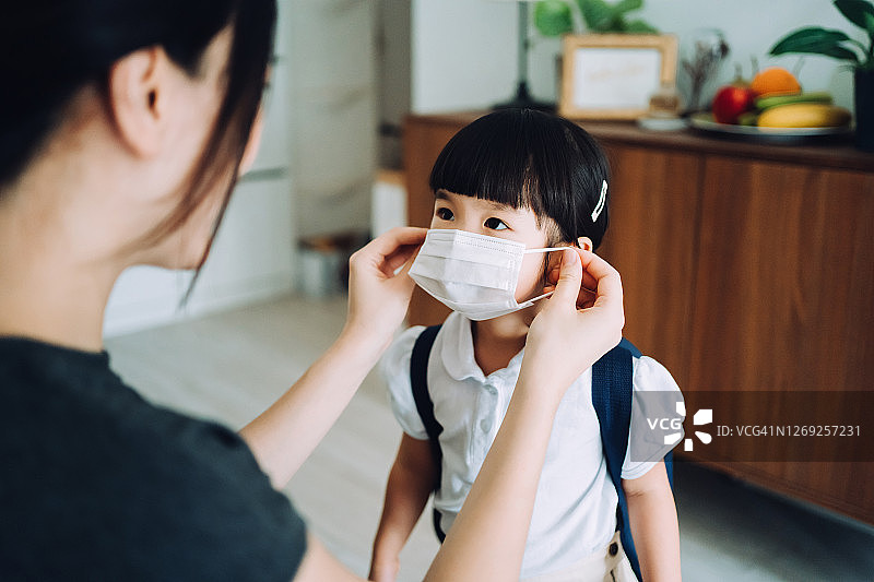 年轻的亚洲母亲为女儿上学的第一天做准备，并为女儿戴上防护口罩，以保护和预防病毒传播图片素材