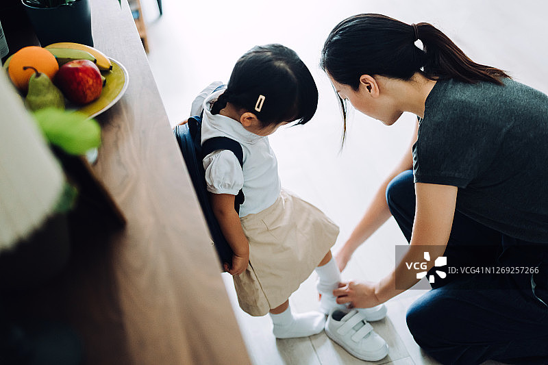 年轻的亚洲母亲在客厅里给她的小女儿穿鞋，为她第一天上学做准备图片素材