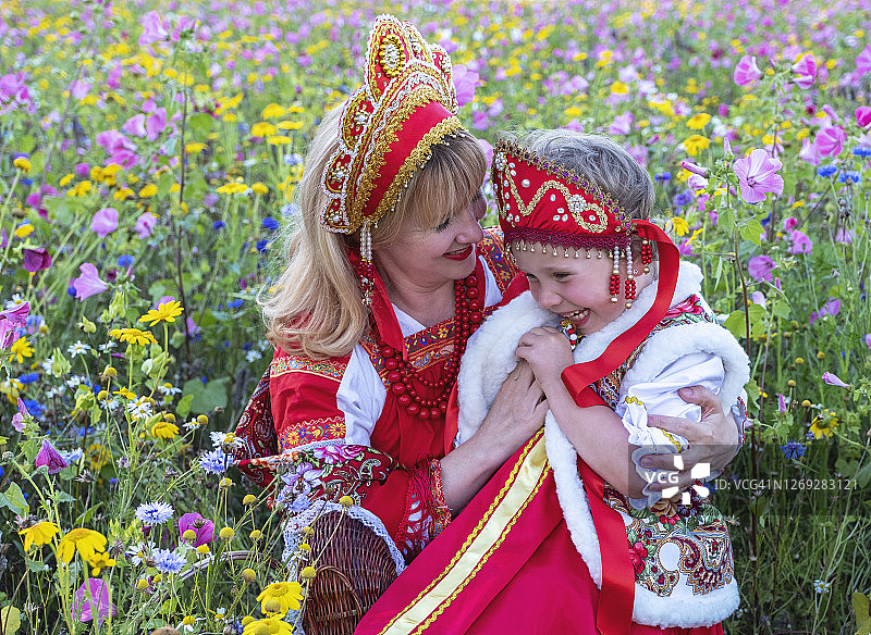 穿着红色连衣裙和戴着科科什尼克头饰的白人妇女和女孩在开满鲜花的草地上。图片素材