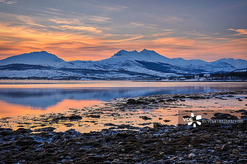 挪威北部特罗姆瑟附近希尤尔斯内斯，一月北极日出，水面倒影，背景是白雪覆盖的山脉和海岸上的小村庄图片素材