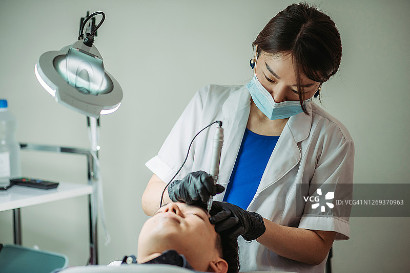 亚洲华裔女美容师在美容院用机器为男士纹眉图片素材