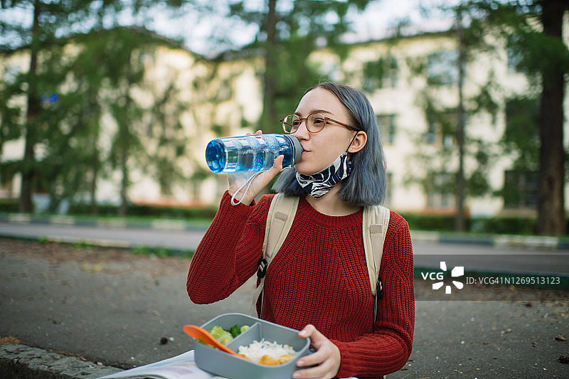 校园里戴可重复使用口罩喝水的少女，新冠隔离后的新常态和返校概念图片素材