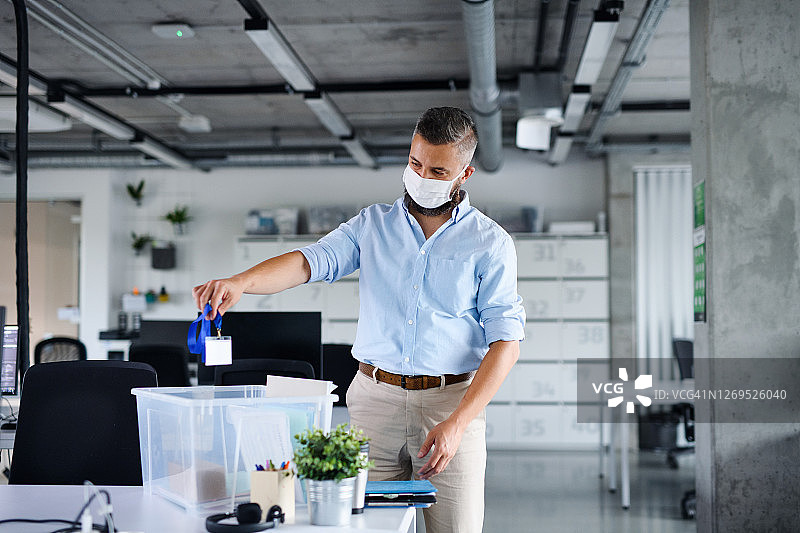 戴着口罩、沮丧的中年男人站在办公桌旁，离开办公室图片素材