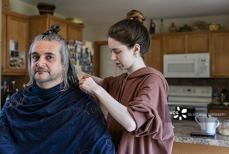 自己动手 - 家庭理发。一个十几岁的女儿给她的父亲，一个50岁留长发的中年男人理发。图片素材