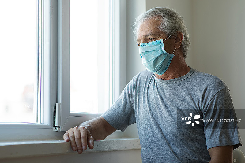 居家隔离、戴着口罩、透过窗户向外看的年长男人图片素材