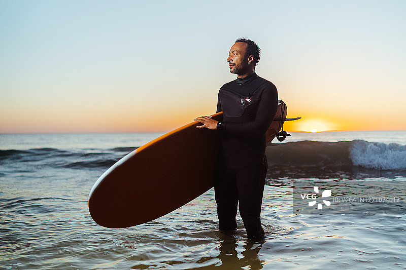穿着潜水衣在日落时分摆姿势的快乐的40岁冲浪男人图片素材