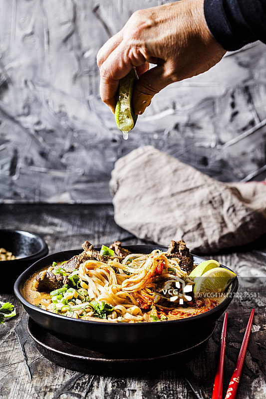 女人将酸橙挤入红咖喱汤中，汤里有米粉、牛肉、蔬菜、葱和花生图片素材