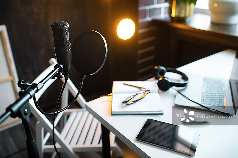 在家播客直播。带有笔记本电脑的音频工作室，麦克风带有防喷罩和耳机，在白色桌子上靠在带有暖色灯的黑墙上。博主概念。图片素材