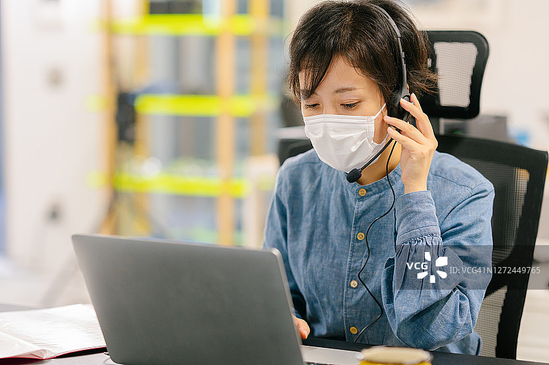 戴防护口罩在办公室使用耳机和笔记本电脑工作的女商人图片素材