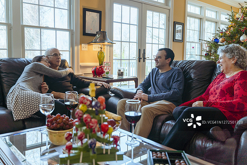 圣诞节家庭团聚，两代人围坐在咖啡桌旁，宽敞的客厅里装饰着巨大的圣诞树，成年女儿拥抱亲吻着年迈的父亲图片素材