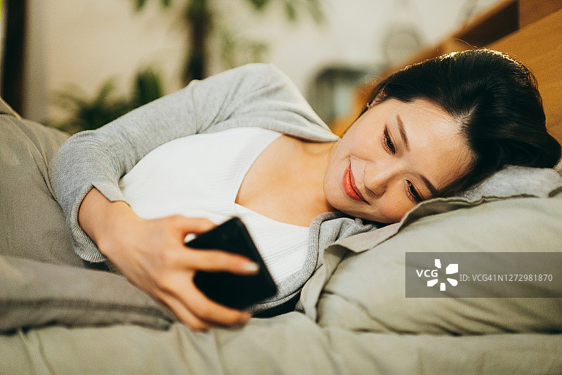 在家中床上使用智能手机，享受轻松夜晚的微笑亚洲年轻女子图片素材