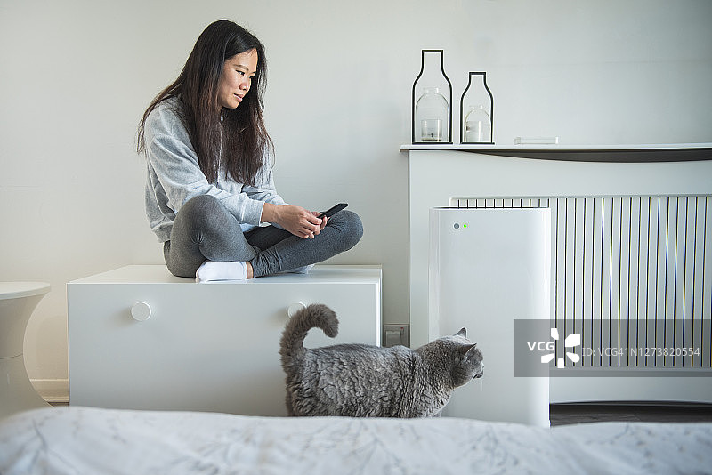 年轻亚洲女性在卧室的储物凳上用手机控制智能空气设备，猫咪走过图片素材
