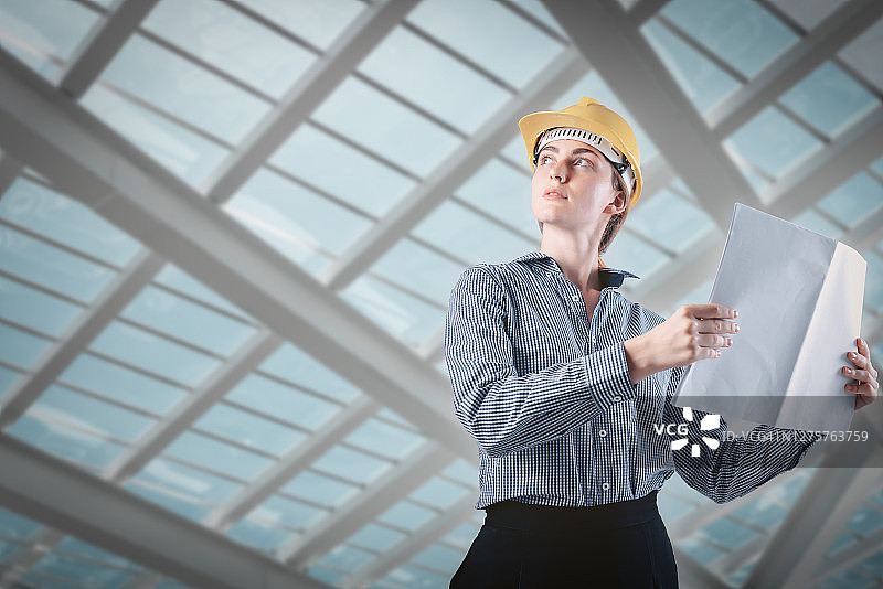 戴着安全帽和安全防护工作服的女土木工程师站在建筑工地上，手持蓝图进行检查。工程职业和建筑行业摄影主题图片素材