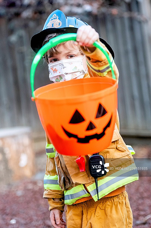 穿着万圣节消防员服装、戴着防护面罩、拿着南瓜灯桶的严肃小男孩看着镜头图片素材