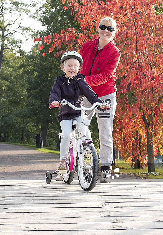 儿童在秋季城市公园骑自行车，年轻女子帮助孩子学习骑自行车图片素材