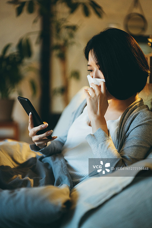 年轻的亚洲女性患有感冒和流感，用纸巾擤鼻涕，同时在床上使用智能手机在家休息图片素材