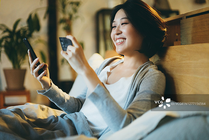 在床上用手机购物的微笑亚洲年轻女性图片素材