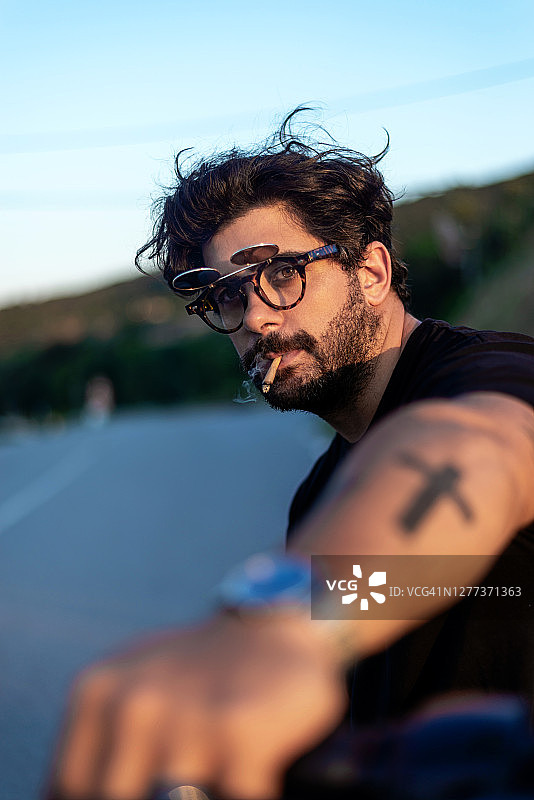 时尚的年轻男子坐在经典的摩托车上在日落时吸烟图片素材