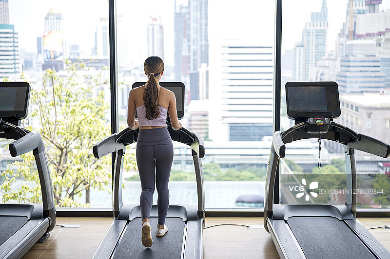 健身女人在健身俱乐部跑步机上进行有氧运动，背景是城市景观的后视图图片素材
