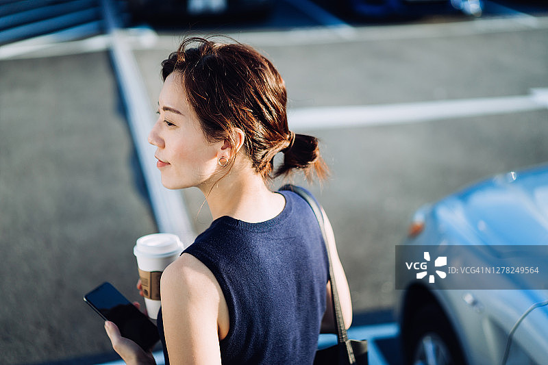 自信的年轻亚洲女商人走出汽车，手持智能手机和咖啡杯。在旅途中的商务概念图片素材