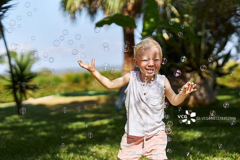 阳光下，一个金发小男孩在花园里吹泡泡，玩得很开心。图片素材